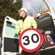 Image of a Worker in a hardhat wearing a hi-viz jacket holding a 30 km per hour sign